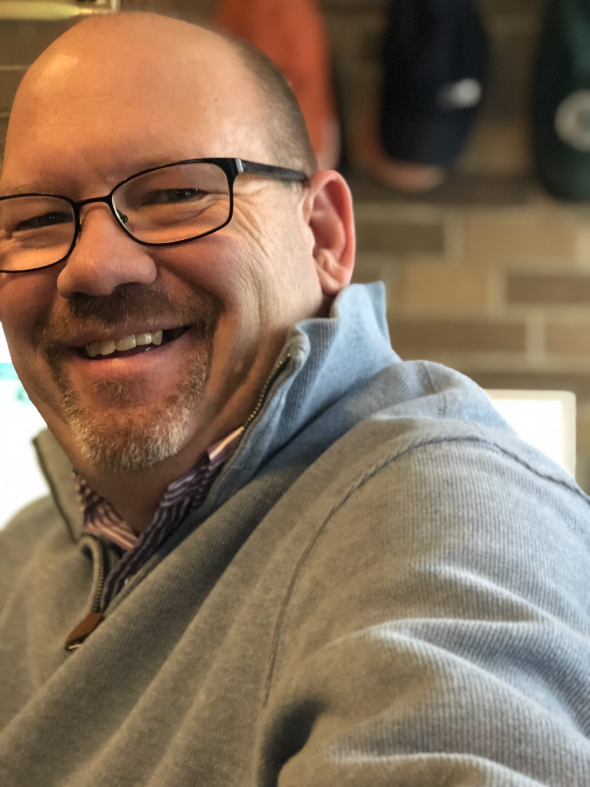 Photo of Jimmy Bornamann, Co-Founder and Chief Digital Operations Officer, smiling warmly, wearing glasses, and a grey pullover, in a cozy indoor setting.