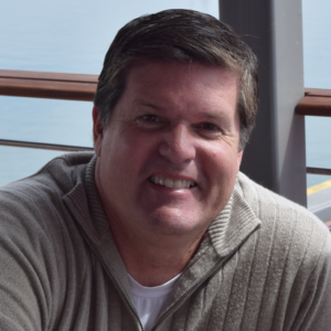 Steve Thompson, Co-Founder & President of Tactiqly, smiles warmly in this photo. Dressed in a casual gray sweater over a white shirt, he's seated with a relaxed, confident posture against a blurred coastal backdrop, embodying a blend of professionalism and approachability.